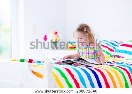 Toddler girl reading a book in bed. Children read books. Kids learning. Preschool kid doing homework in bedroom. Colorful textile bedding for child and baby room. Preschoolers learn and study at home