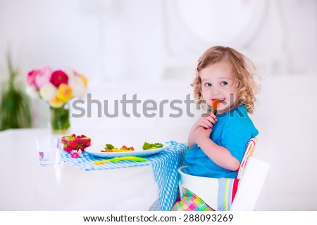 Child having vegetables for lunch. Healthy fruit and vegetable meal for children. Kids eat in a white dining room or kitchen. Toddler kid having breakfast. Food for preschooler at home or daycare.