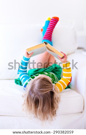 Little girl reading a book relaxing on a white couch. Kids read books at home or preschool. Children learning and doing homework after school. Child playing. Toddler kid in colorful dress on a sofa.