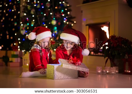 Family on Christmas eve at fireplace. Kids opening Xmas presents. Children under Christmas tree with gift boxes. Decorated living room with traditional fire place. Cozy warm winter evening at home.
