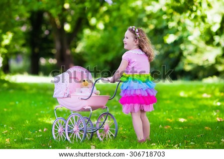 Little girl pushing toy stroller with bear. Toddler kid in pink dress playing with doll buggy. Kids birthday party. Children play outdoors. Mother and baby role game. Family summer fun. Preschool toys