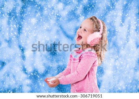 Little girl in pink knitted sweater and fur earwarmer catching snowflakes in winter park. Kids play outdoor in snowy forest. Children catch snow flakes. Toddler kid playing outside in snow storm.