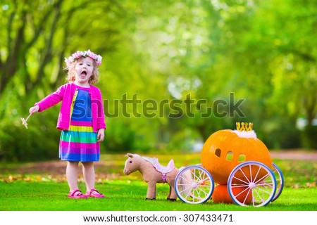 Cute curly little girl playing Cinderella fairy tale holding magic wand next to a pumpkin carriage in autumn park at Halloween. Kids trick or treat at pumpkin patch. Family with children carving.