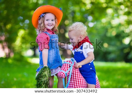Little boy and girl dressed up as cowboy and cowgirl playing with toy rocking horse in park. Kids play outdoors. Children in Halloween costumes at trick or treat. Toys for preschooler or toddler child