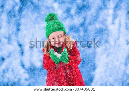 Little girl in red jacket and green knitted hat catching snowflakes in winter park on Christmas eve. Kids play outdoor in snowy winter forest. Children catch snow flakes on Xmas. Toddler kid playing.