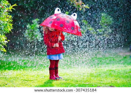Little girl with red umbrella playing in the rain. Kids play outdoors by rainy weather in fall. Autumn outdoor fun for children. Toddler kid in raincoat and boots walking in the garden. Summer shower.
