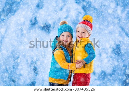 Little girl and boy in yellow and blue knitted hat catching snowflakes in winter park on Christmas eve. Kids play outdoor in snowy winter forest. Children catch snow flake on Xmas. Toddler kid playing