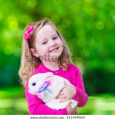 Children play with real rabbit. Laughing child at Easter egg hunt with white pet bunny. Little toddler girl playing with animal in the garden. Summer outdoor fun for kids with pets.