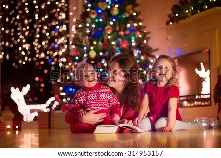 Family on Christmas eve at fireplace. Mother and little kids opening Xmas presents. Children with gift boxes. Living room with traditional fire place and decorated tree. Cozy winter evening at home.