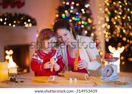 Mother and little girl baking Christmas pastry. Children bake gingerbread. Toddler child preparing cookie for family dinner on Xmas eve. Decorated kitchen or dining room with fireplace, tree, candles.