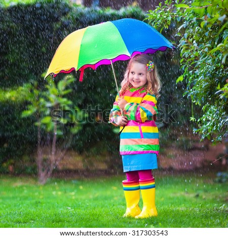 Little girl with colorful umbrella playing in the rain. Kids play outdoors by rainy weather in fall. Autumn fun for children. Toddler kid in raincoat and boots walking in the garden. Summer shower.