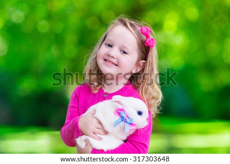 Children play with real rabbit. Laughing cute child at Easter egg hunt with white pet bunny. Little toddler girl playing with animal in the garden. Summer outdoor fun for kids with pets.