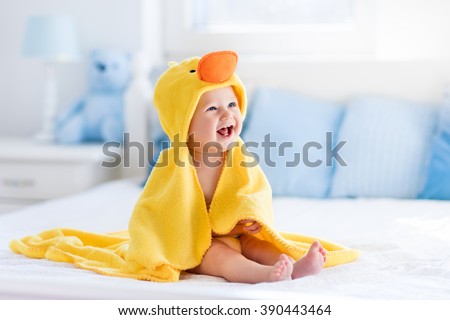 Happy laughing baby wearing yellow hooded duck towel sitting on parents bed after bath or shower. Clean dry child in bedroom. Bathing and washing of little kids. Children hygiene. Textile for infants. 商業照片 © 