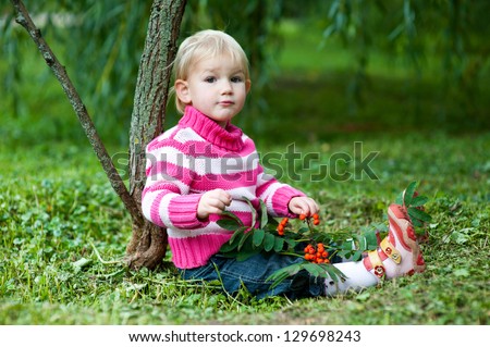 Funny blond infant girl sitting under tree in green forest and playing with berries