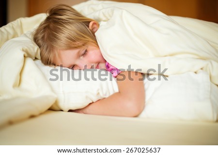 Happy little girl sleep on bed under blanket and posing in room of her flat holding a pillow in hands