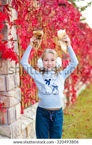 Very beautiful emotional model toddler girl stay at colorful autumn street and enjoy chocolate bars. Red plants on background.