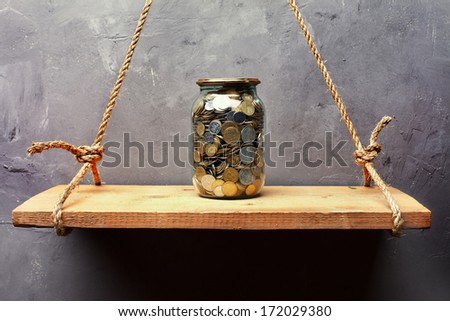 Glass jar with coins on the old wood shelf hanging on rope on textured wall background