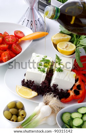 Mediterranean salad with tomatoes, olives, cheese and a lemon, served on a white table, with olive oil pouring on a peaces of cheese