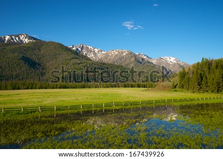 Hills, peaks, and ridges of the Rocky Mountains reflected in the water of a lake bordering a farm.