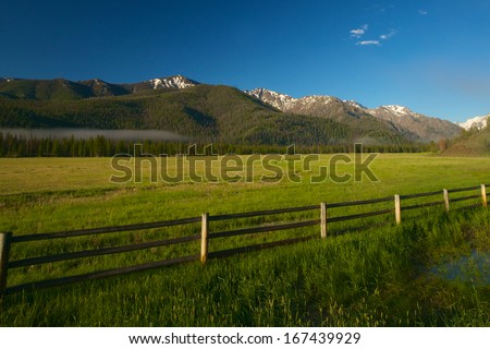 Wooden rail fence marks the border of a farm beneath a  range of snow-capped mountains.