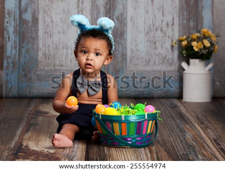 Adorable baby boy sitting on a rustic wood floor, playing with an Easter Basket full of plastic eggs and wearing blue bunny ears.