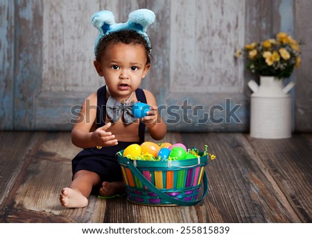 Adorable baby boy sitting on a rustic wood floor, playing with an Easter Basket full of plastic eggs and wearing blue bunny ears.