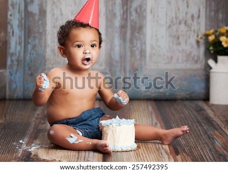 Adorable baby boy, sitting on a wood floor, wearing a party hat and eating a small cake.  Room for your text.