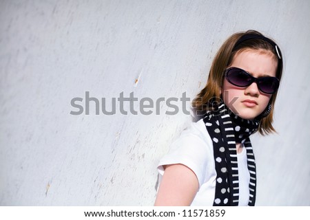 Head and shoulders portrait of a pretty preteen in sunglasses against a white, sunlit wall.