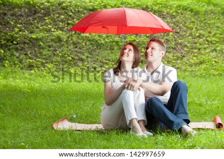 Beautiful young couple in love sitting on green summer meadow under red umbrella
