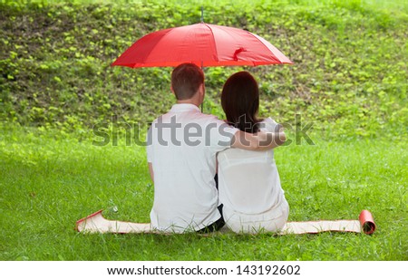 Couple in love sitting on green summer meadow under red umbrella