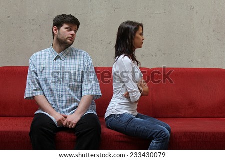 Sad couple sitting on a red sofa - young man hopefully watching his girlfriend while waiting for her forgiveness
