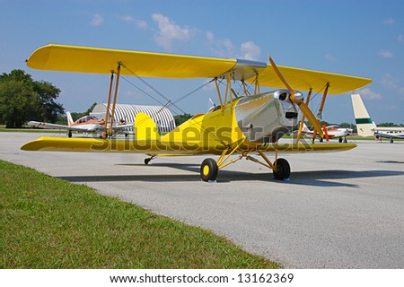 A bright yellow, vintage biplane parked on the tarmac at an air show with other planes in the background.