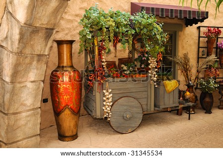 A rustic wooden cart filled with fruits and vegetables in the marketplace with a huge vase in the foreground.