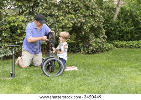 Father and Son, 6 year old boy, are spending quality time together fixing a bike. They are in a backyard sitting on grass, trees behind them with a bicycle between them