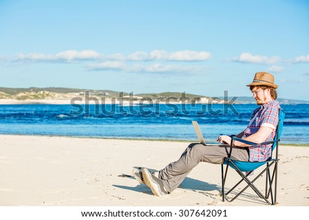 Happy young man in a straw hat sitting in the beach chair near the ocean working on his laptop