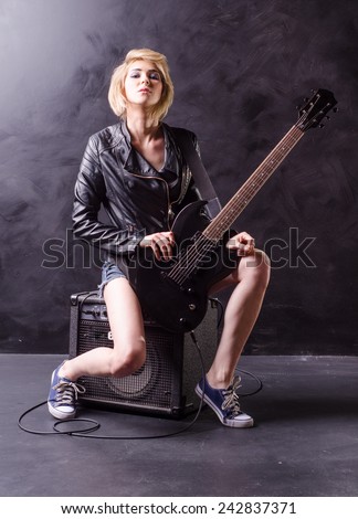Beautiful young blonde siting on the amp dressed in black leather jacket with electric guitar on a black background