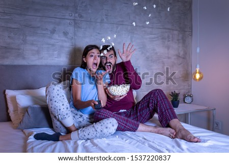 Beautiful young couple in love, sitting in bed side by side, eating popcorn and having fun watching a horror movie at night Stock fotó © 