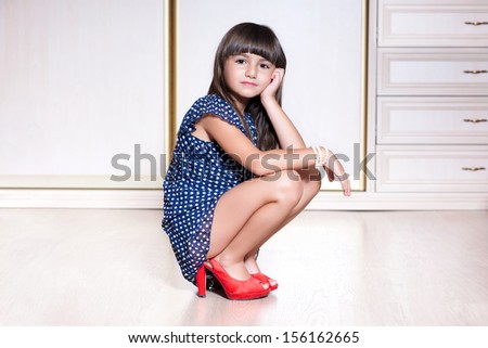 Little beautiful girl in a blue dress and red shoes sitting in a bright room