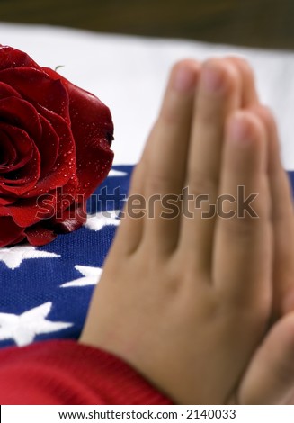 A young child prays for a fallen loved one. In the background is a folded flag and red rose.
