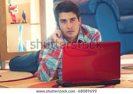 Portrait of handsome young man lying on the carpet floor with laptop while listening to phone