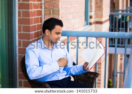 Closeup portrait of young happy handsome man student worker employee sitting and reading enjoying book journal, drinking beverage on outside balcony, isolated on apartment background. Positive emotion