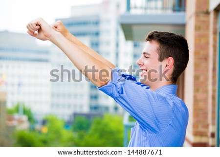 Portrait of happy, handsome, successful guy raising hands, arms outstretched in celebration and success, enjoying life on outside balcony, isolated on city background with trees and buildings