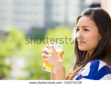 A portrait of a beautiful young female relaxing on a balcony on a sunny summer day, drinking orange juice, in her new apartment, background of a city scenery and green trees. Urban lifestyle.