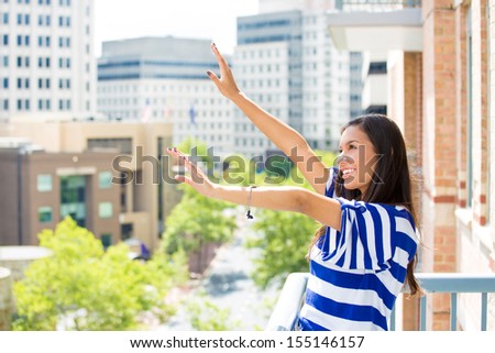 A portrait of a beautiful young female relaxing on a balcony on a sunny summer day, enjoying her weekend, in her new apartment, background of a city scenery and green trees. Urban lifestyle.