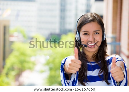 Closeup portrait of young female customer service representative, call center agent, support staff or operator with phone headset on balcony giving us thumbs up, isolated on a city background