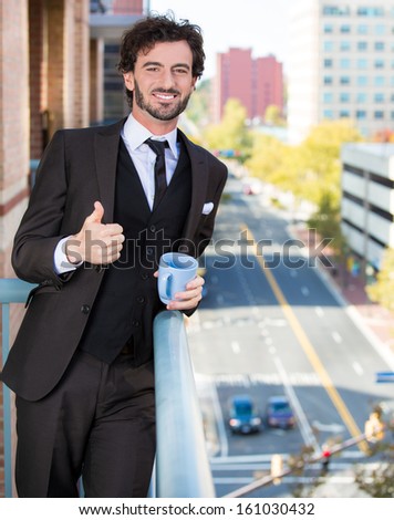 Closeup portrait of good looking smiling, happy businessman giving you a thumbs up, enjoying a drink, standing on his balcony, isolated on a city background. Urban life style, living of an executive