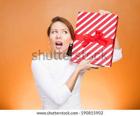 Closeup portrait happy, excited young woman about to unwrap red birthday gift box, wondering what is inside isolated orange background. Positive emotion, facial expression, feeling, attitude, reaction