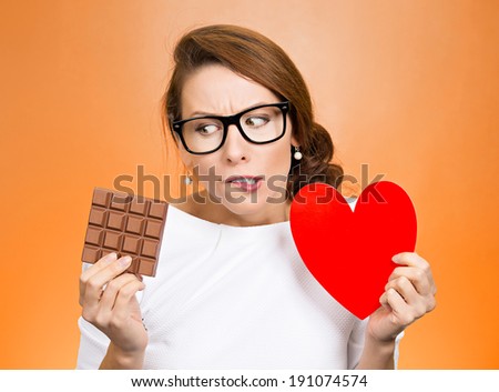 Closeup portrait, headshot beautiful, young nerd woman with black glasses holding heart craving square milk chocolate isolated orange background. Food diet option dilemma. Sweet temptation. Expression
