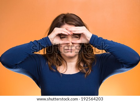 Closeup portrait young, curious funny woman, looking through fingers like binoculars, searching something, unhappy, disgusted with what waiting in future, isolated orange background. Facial expression