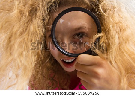Closeup portrait beautiful, surprised little girl with curly blonde hair looking through magnifying glass. Human emotions, facial expressions, attitude, reaction, curiosity, life perception, childhood